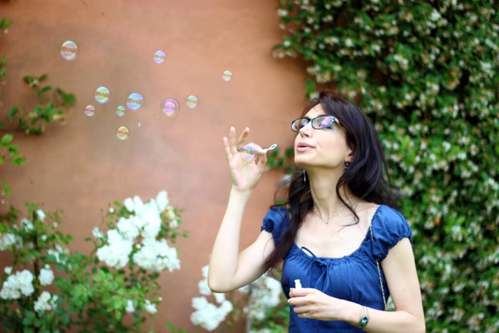valentina antonaci soffia bolle di sapone. indossa un vestitino blu notte. sullo sfondo dei fiori